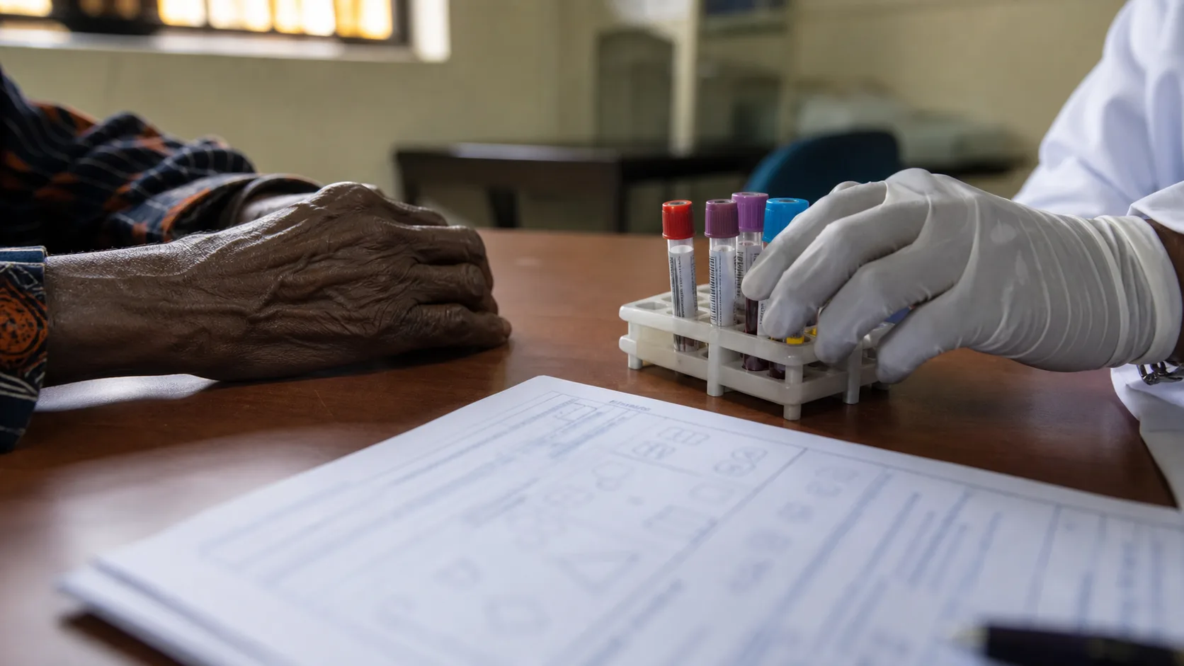 An older adult's hand resting on a clinic table beside blood-sample tubes held by a gloved clinician